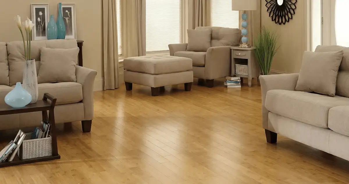 A living room with professionally installed bamboo flooring, beige sofas, a matching ottoman, a side table with a blue lamp, vases, framed pictures, and large windows with blinds and curtains in South Bend, IN